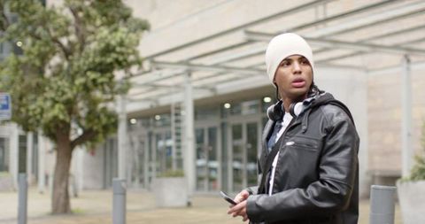 Urban commuter wearing beanie headphones standing in plaza looking right holding smartphone