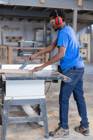 Carpenter cutting lumber on table saw wearing safety gear in woodworking workshop