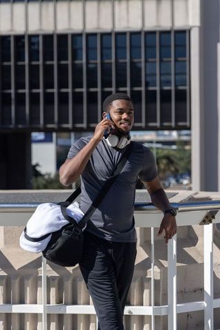 African american man leaning on railing talking on phone with duffel and headphones
