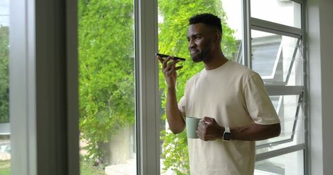 Man standing by window talking on smartphone holding coffee cup in bright modern interior