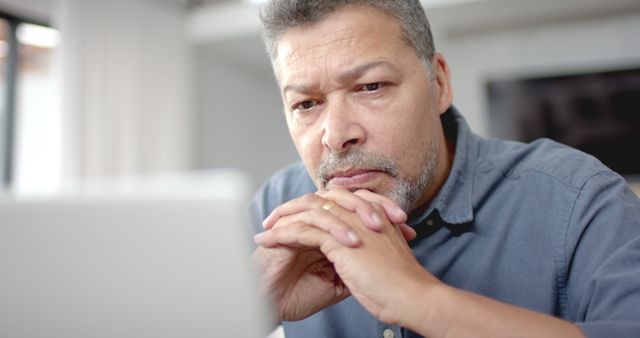 Older man with gray hair focusing on laptop screen in his home office. He is wearing a blue shirt and has a serious expression. This image can be used to depict remote work, focused tasks, technology use among older adults, digital literacy, or home office scenarios.