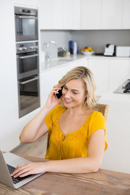 Woman talking on mobile phone while using laptop in kitchen at home