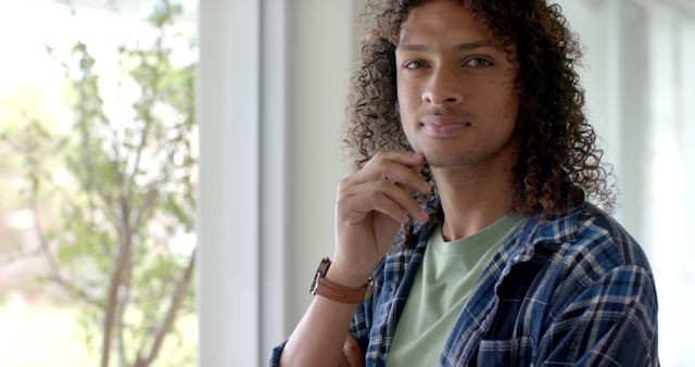 Young man with curly hair pensive by window. Wearing casual, plaid shirt over T-shirt, thoughtful expression. Ideal for themes of introspection, daily life, casual attire, modern youth. Perfect for use in lifestyle blogs, mental health articles, or fashion advertising.