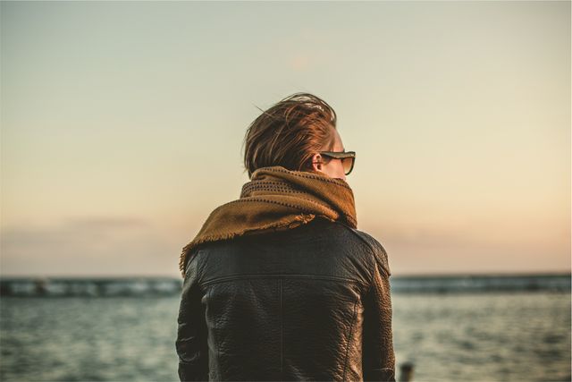 Person wearing a leather jacket and scarf is peacefully enjoying the view of the sea during sunset. Perfect for use in advertisements, travel blogs, relaxation themes, lifestyle magazines, and scenic backgrounds.