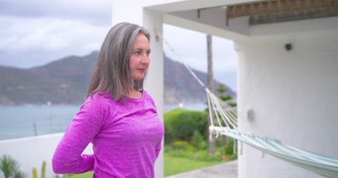 Mature woman standing on coastal terrace in magenta top with hammock and ocean bay