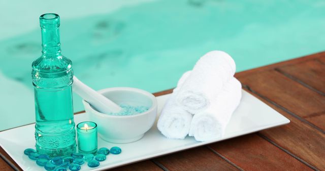 This scene shows neatly rolled spa towels on a white tray alongside a blue glass bottle, a flame-lit tealight candle, bath salts in a white mortar and pestle, and decorative blue glass pebbles. Perfect for use in marketing material for a luxury spa, wellness retreats, relaxation therapies, or beauty and skincare products.