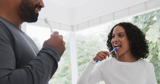 A couple is brushing their teeth together in a bright, spacious bathroom. They seem joyful as they engage in this daily morning routine. This image can be used for dental care advertisements, promoting healthy hygiene habits, or for relationship and family-related content.