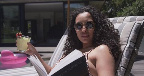 African american woman relaxing poolside reading book and sipping cocktail with sunglasses