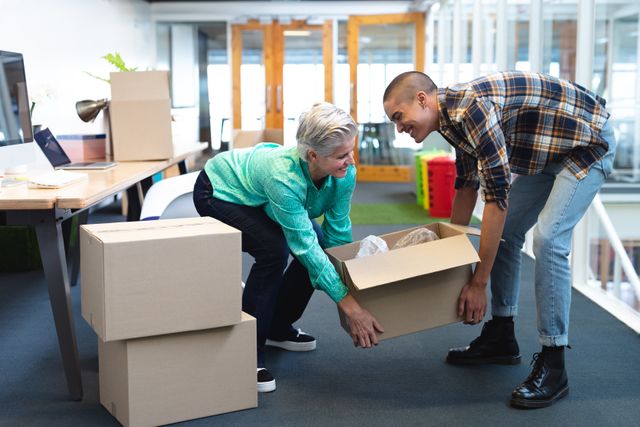 Diverse male and female colleagues are seen working together to move cardboard boxes in a modern office environment. This image is ideal for illustrating teamwork, collaboration, and the dynamic nature of startup businesses. It can be used in articles or advertisements related to office relocation, team building, or startup culture.