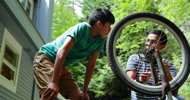 A father and his son repairing a bicycle together outside their home during summer. This moment captures the essence of family bonding and teamwork. Ideal for use in articles, ads, or blogs focusing on family activities, learning new skills together, or promoting healthy outdoor hobbies.