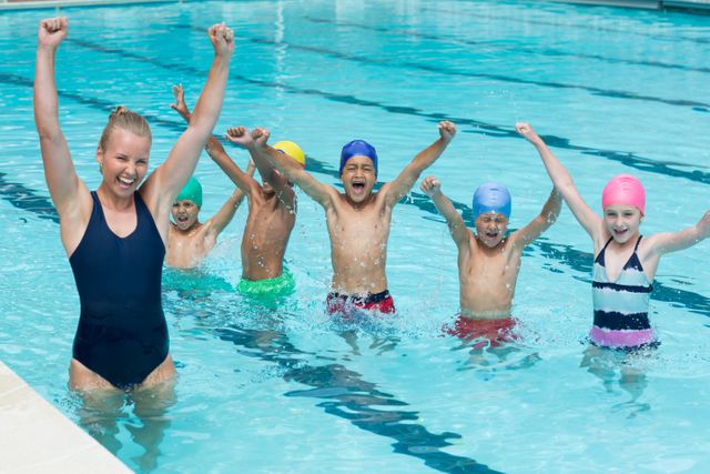 Group of children with a cheerful female instructor in a swimming pool, all raising their arms in excitement. Ideal for use in advertisements for swimming lessons, summer camps, children's activities, or fitness programs. Highlights fun and learning in a safe aquatic environment.