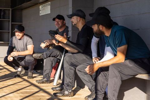 Sunlit dugout team bonding five baseball players sitting chatting holding bat glove ball