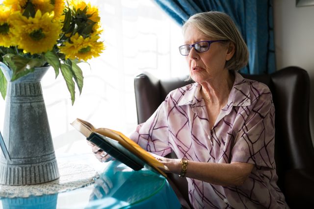 Senior woman wearing glasses reading a book while sitting on an armchair in a well-lit living room. Sunlight streams through the window, illuminating the room. A vase of sunflowers adds a touch of color to the scene. Ideal for use in articles about retirement, elderly care, leisure activities for seniors, or home decor.