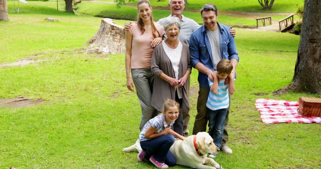 Multigenerational family standing together and smiling at a park, enjoying a relaxed picnic with their dog. Suitable for use in lifestyle, family-oriented, and leisure activity promotions. Can be used in advertisements focused on outdoor activities, family quality time, or pet-friendly environments.