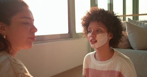 Diverse women sitting on floor, chatting and enjoying facial mask self-care in sunlit room
