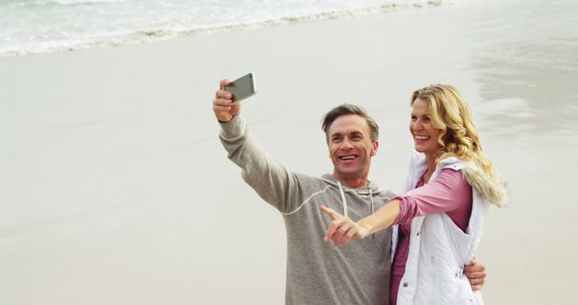 Mature couple standing on sandy beach taking selfie with smartphone. They look happy and relaxed, enjoying their time together with the ocean in the background. Ideal for use in travel advertisements, social media posts about relationships or outdoor activities, and health and wellness campaigns.
