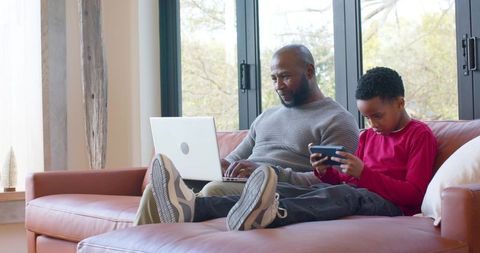 African American Father and Son Relaxing on Sofa Using Laptop and Handheld Console