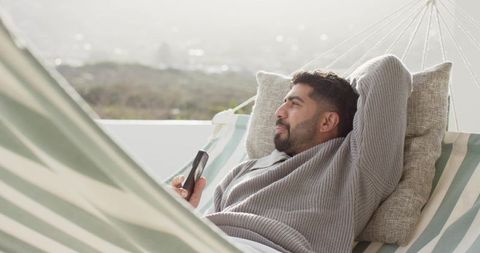 Indian man relaxing in striped hammock on rooftop terrace holding smartphone, serene