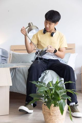 Young asian man unpacking and inspecting metal desk lamp on bed in minimalist bedroom