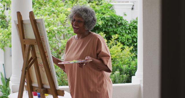 An elderly woman with gray curly hair is happily painting on a canvas in an outdoor studio with greenery in the background. She is holding a palette and appears to be deeply engrossed in her work. This image can be used to promote senior activities, artistic hobbies, and creative environments. Ideal for websites and articles focused on senior living, mental health, and lifestyle.