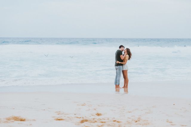 A young couple stands on a sandy beach in a loving embrace with the ocean waves crashing behind them. This scene can be used for romantic getaway promotions, beach lifestyle blogs, relationship advice content, or travel brochures featuring seaside destinations.