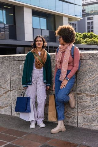 Two women standing and talking on urban sidewalk with shopping bags and casual streetwear