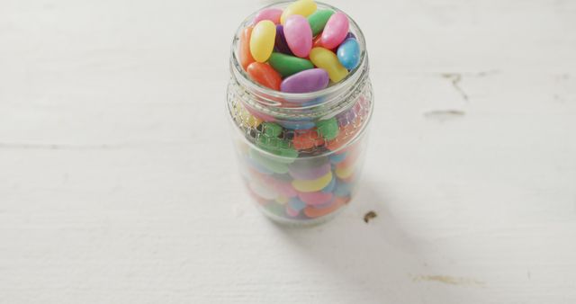 Brightly colored jelly beans filling a clear glass jar on a white table. Ideal for use in content relating to food, desserts, snacks, candy, sweets, holidays, parties, celebrations, childhood, and confectionery products. Useful for advertisements, blog posts about candy or treats, and children's parties visuals.