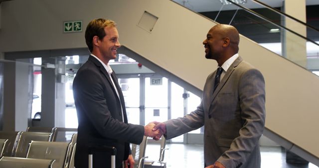 Image depicting two businessmen shaking hands and smiling in a modern office lobby. This can be used for portraying positive business interactions, partnerships, professional greetings, corporate environment, and business success stories.