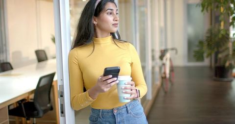 Indian woman leaning in modern glass office corridor holding smartphone and coffee cup