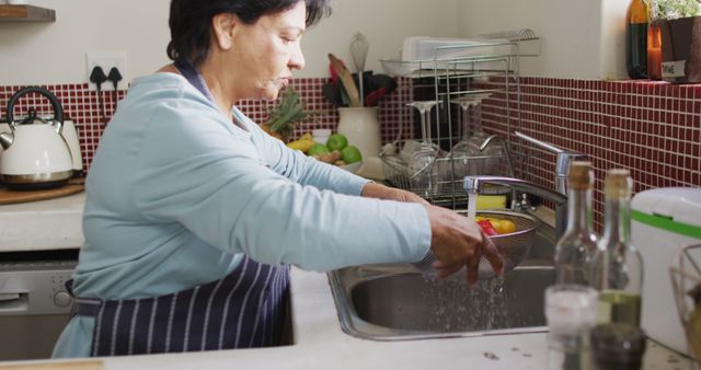 Senior woman washing vegetables in kitchen sink. This image is perfect for illustrating healthy lifestyles, home activities, or cooking scenes featuring senior citizens. It can be used in articles related to health and wellness, senior living, or culinary topics.