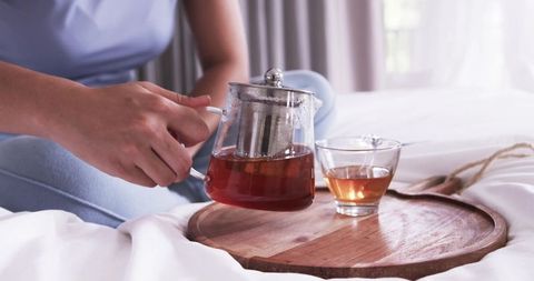 Morning ritual pouring herbal tea from glass teapot on wooden tray over white linens