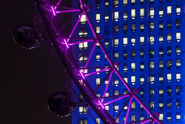 A neon-lit Ferris wheel standing out against a blue-lit skyscraper at night. Useful for themes involving city nightlife, modern architecture, urban activities, and vibrant cityscapes. Ideal for travel, tourism, and advertising related content.
