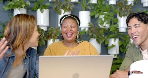 Diverse team laughing and collaborating around laptop with plant wall for modern coworking