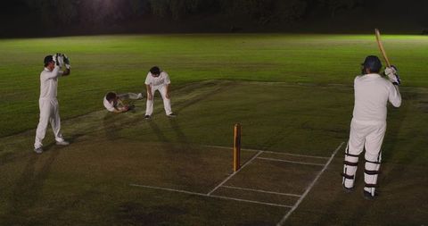 Night cricket with asian players batting and fielding under floodlights on grass pitch
