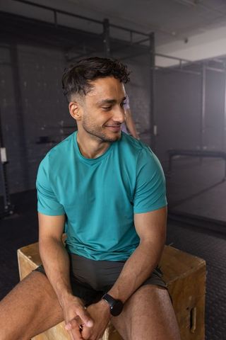 South asian man sitting on plyometric box checking smartwatch in gym training area