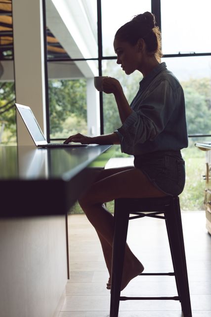 Side view of woman drinking coffee while using laptop on worktop in kitchen at comfortable home