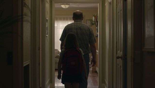 Parent and child walking down home hallway toward sunlit living room with red backpack