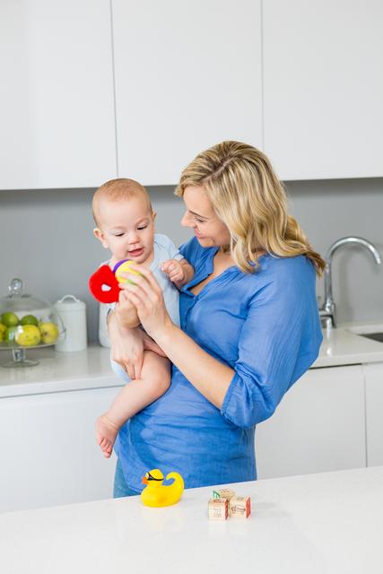 A mother holding her baby boy in a modern kitchen, engaging with him using toys. They are smiling and bonding, suggesting themes of family, love, and domestic life. The clean and bright kitchen background emphasizes a warm home environment. Ideal for use in advertisements, parenting blogs, family-centric articles, and home decor promotions.