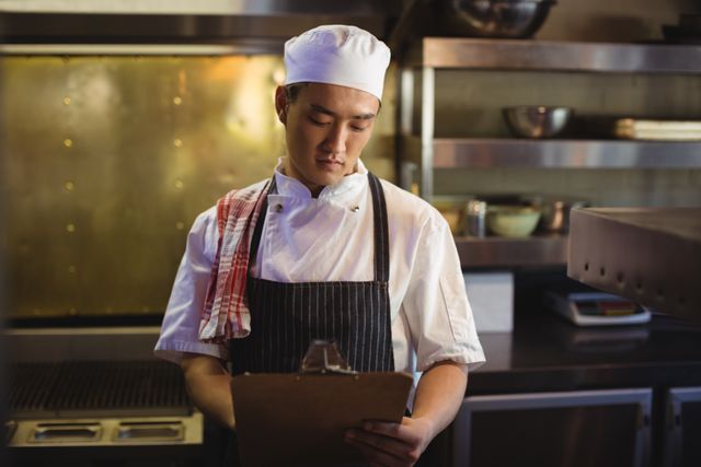 Chef in a commercial kitchen writing on a clipboard, ideal for illustrating professional kitchen environments, culinary arts, restaurant management, and food industry operations.