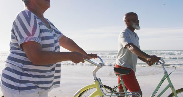 Smiling senior african american couple walking with bicycles at the beach. healthy outdoor leisure time by the sea.
