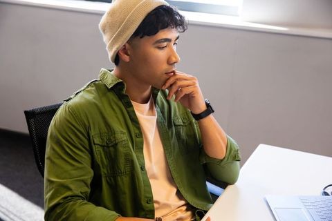 Contemplative asian man in beanie and smartwatch working at laptop in modern office