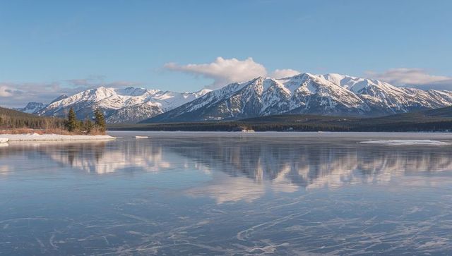 Snow-capped mountains reflecting on frozen alpine lake with evergreen peninsula at sunrise
