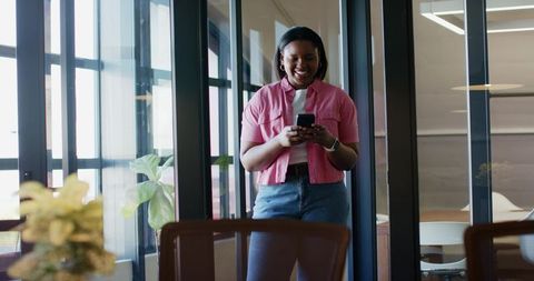 African american woman standing near glass wall smiling using smartphone in modern office