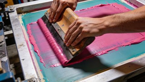 Artisan screen printing hands spreading magenta ink with wooden squeegee on teal mesh