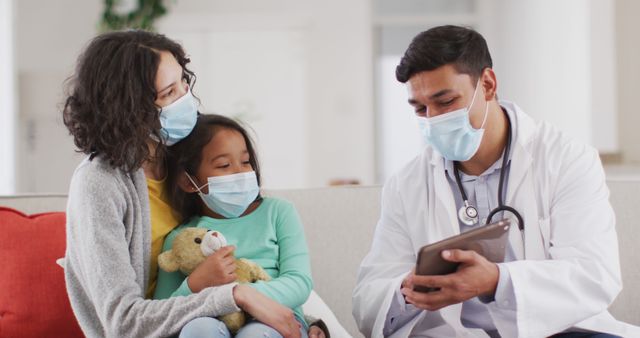 Hispanic male doctor talking to mother and daughter at home, all wearing face masks. medical professional making patient home visit during coronavirus covid 19 pandemic.