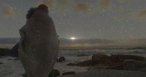 Woman wrapped in fur-trimmed blanket watching sunrise over rocky coastline