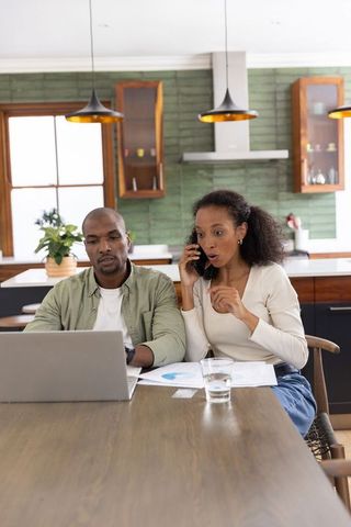 Diverse couple reviewing finances at kitchen table taking phone call and checking charts