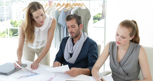Three fashion designers working together in a well-lit studio space. Concepts like collaboration, creativity, and teamwork can be highlighted using this image. Ideal for articles and promotions related to fashion design, teamwork in creative professions, and office work environments.