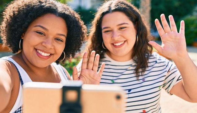 Two friends waving during video call in sunny backyard smiling at smartphone on tripod
