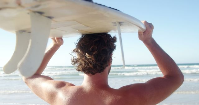 A young Caucasian man carries a surfboard on his head as he walks towards the ocean, with copy space. His anticipation for an exciting surf session is palpable against the backdrop of the inviting sea.